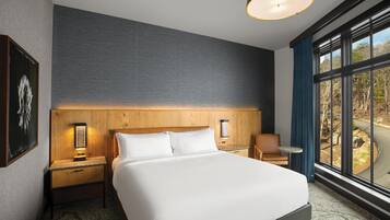 Une chambre d’hôtel avec un grand lit, une table de chevet, une petite table, une chaise et une grande fenêtre donnant sur une route.