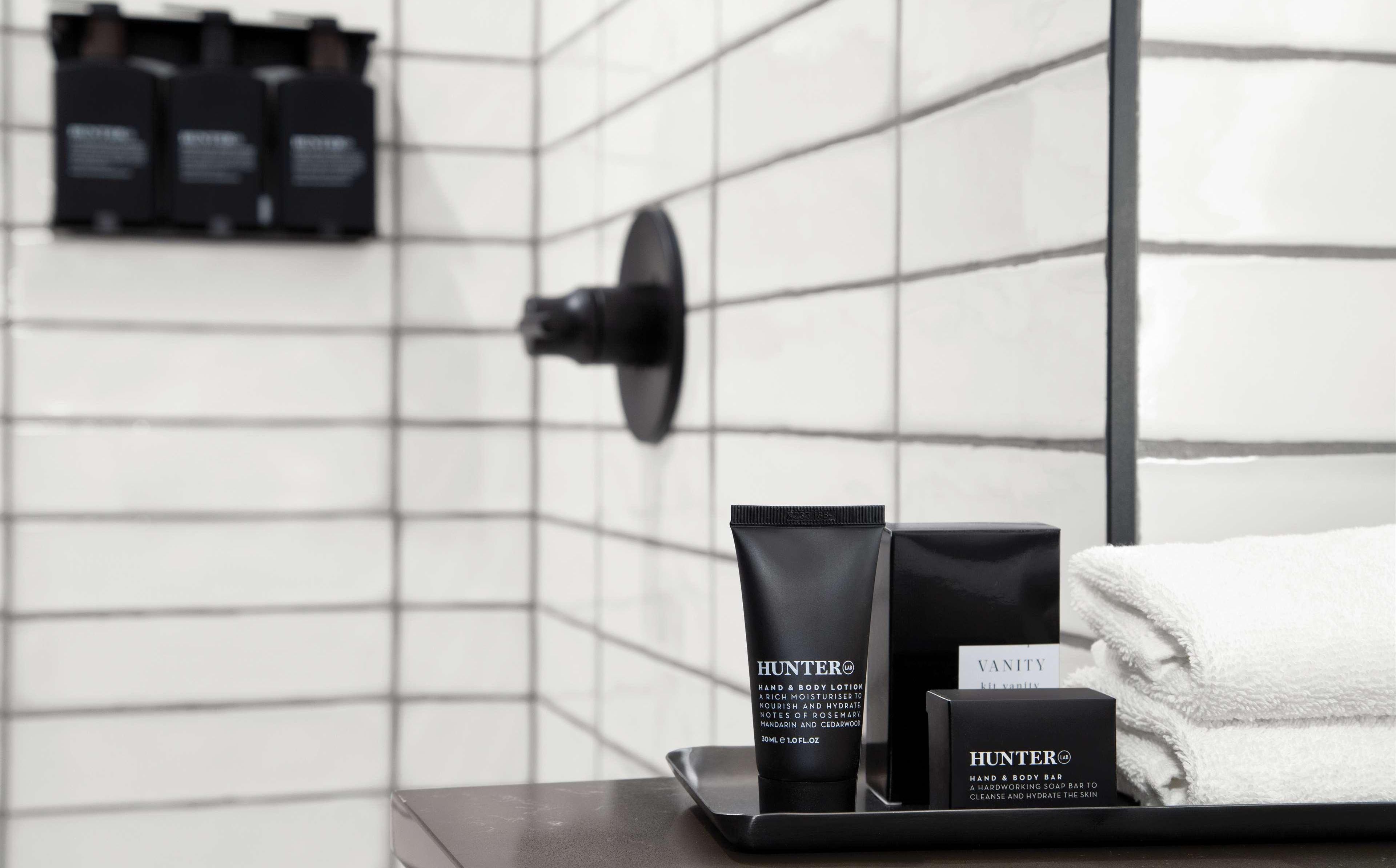 A bathroom with a white tiled wall, a black hand and body lotion bottle, a bar of soap, and a white towel on a tray.