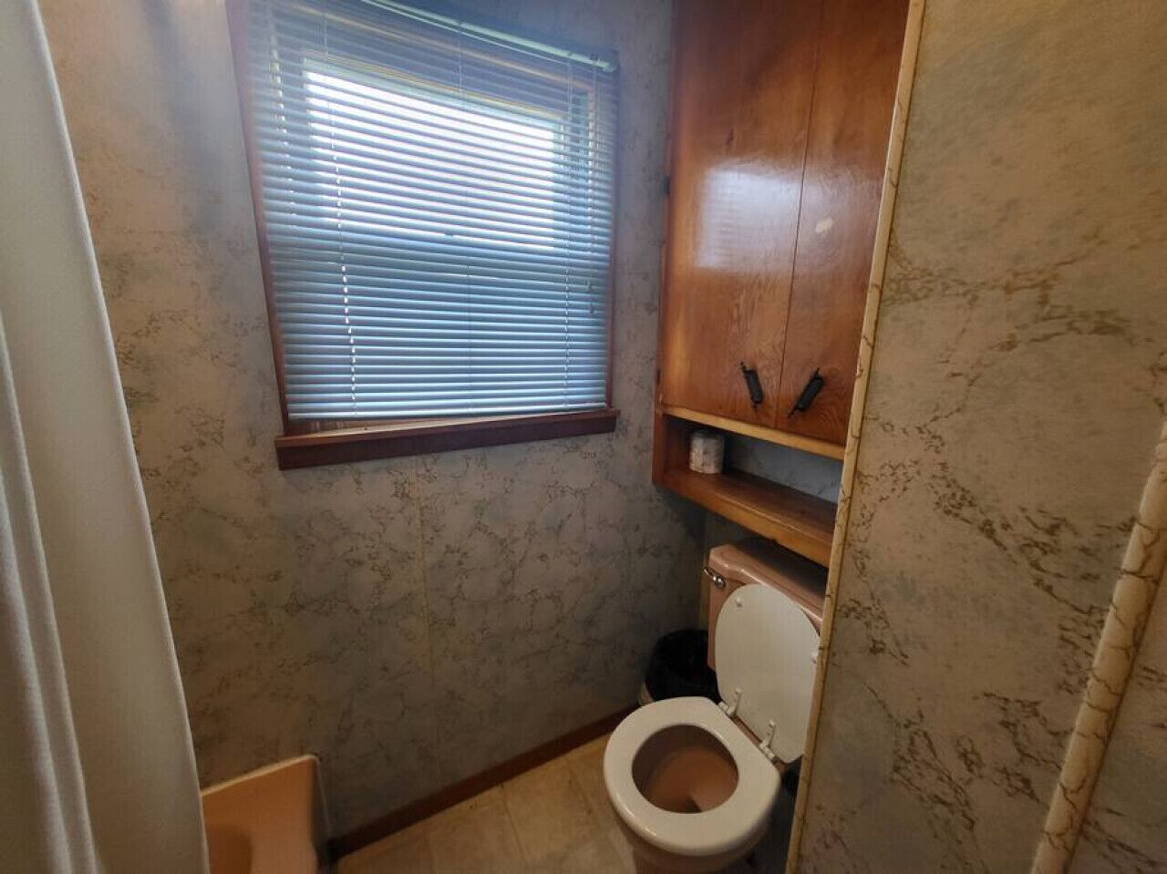 Un baño pequeño con inodoro, una ventana con persianas y un armario de madera sobre el inodoro.