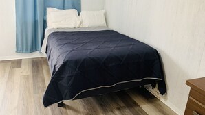 A bedroom with a bed, white pillows, a dark blue bedspread, light wooden flooring, and a blue curtain.