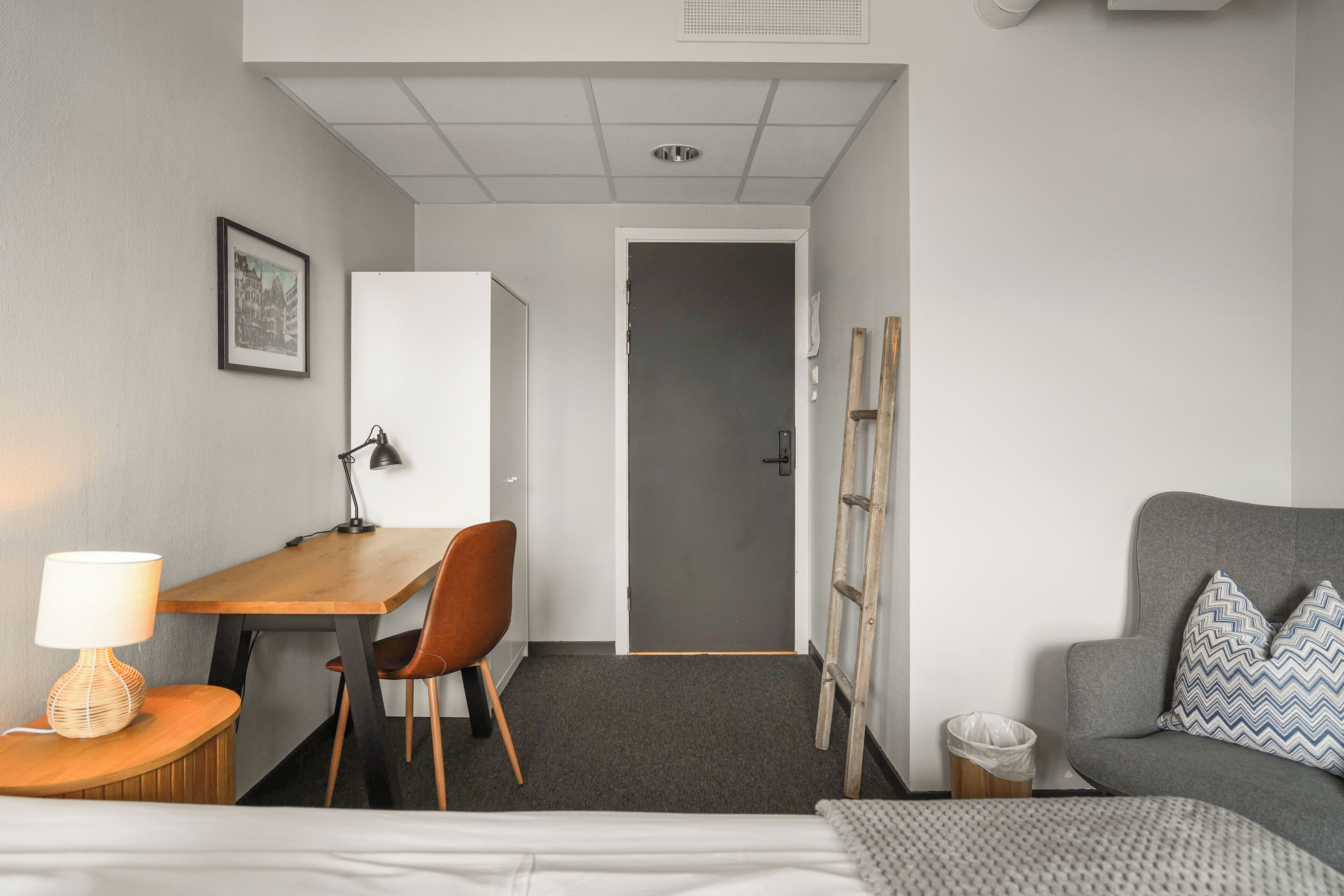 A hotel room with a bed, a desk with a chair, a sofa, a lamp, and a picture on the wall.