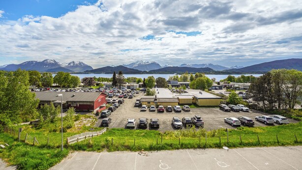 Un parking rempli de voitures, un bâtiment avec un toit rouge et des montagnes en arrière-plan.