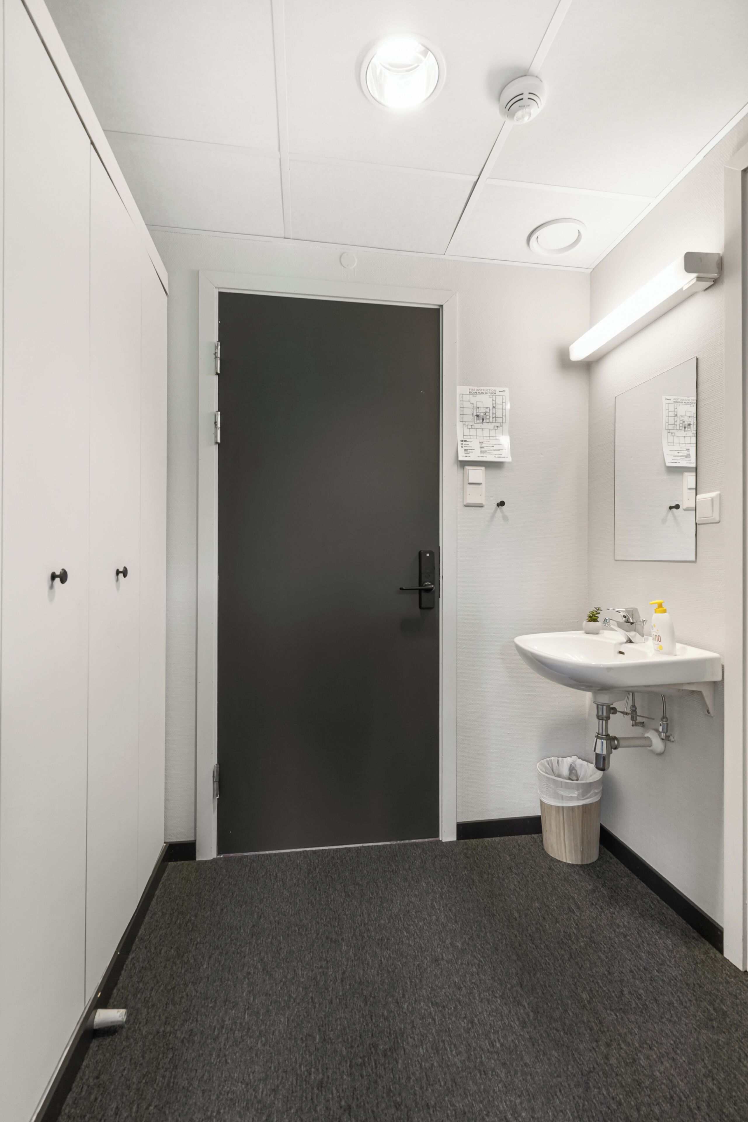 A bathroom with a white sink, a mirror, a trash can, and a door.