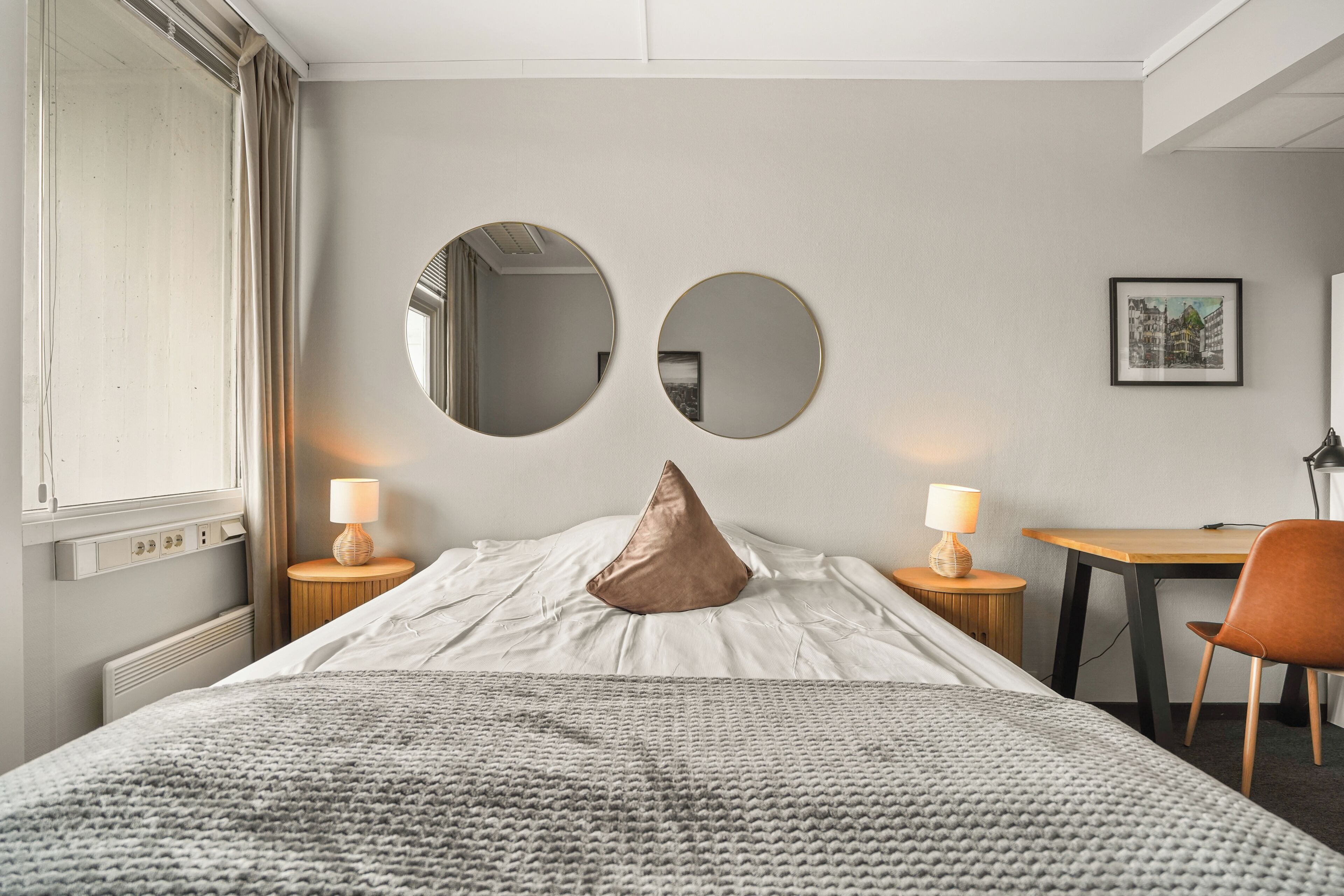 A hotel room with a bed, two bedside tables with lamps, a round mirror, a pillow, a framed picture, and a desk with a chair.