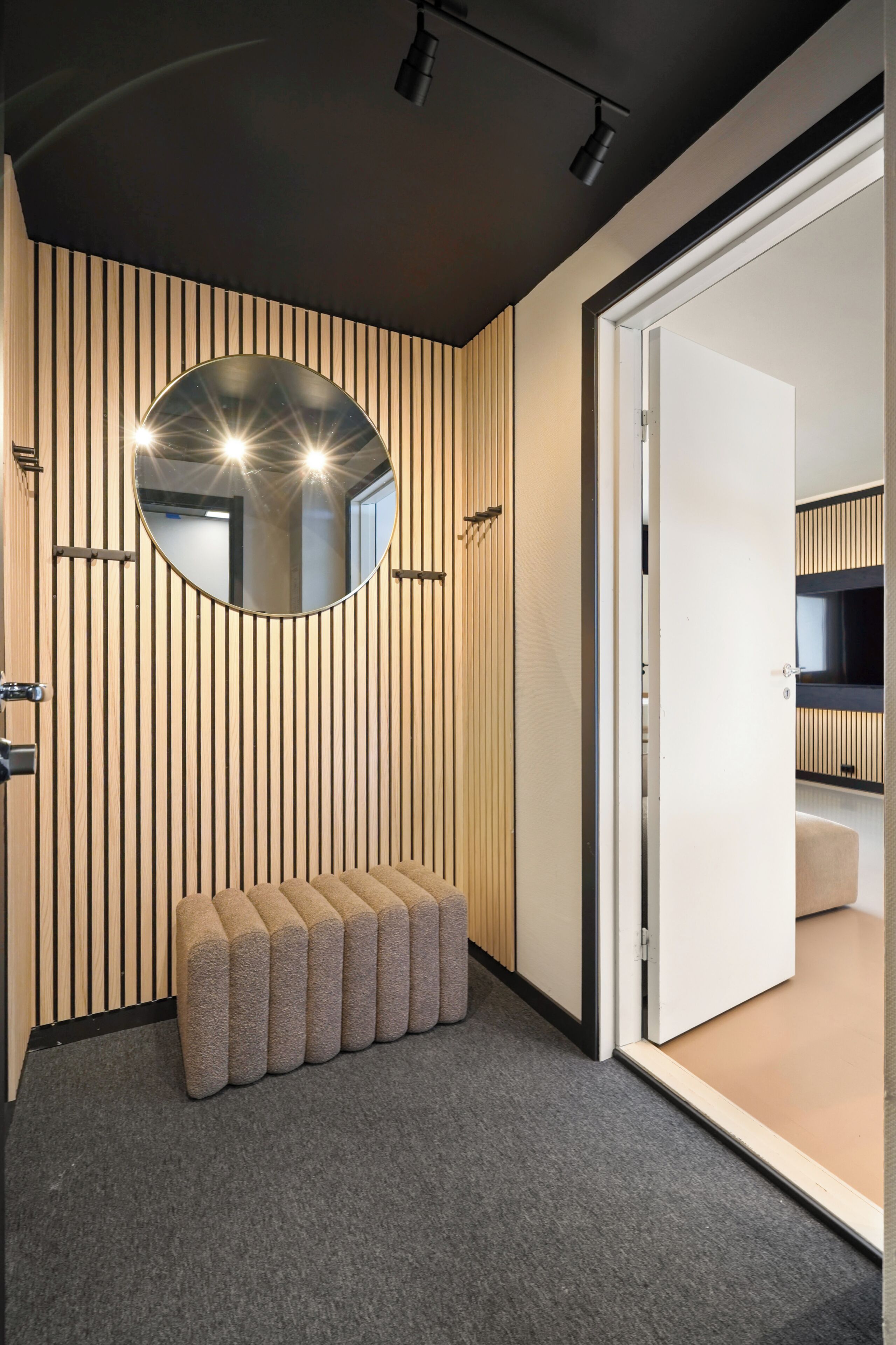 A modern room with a wooden slat wall, a round mirror, a bench, and a door leading to another room.