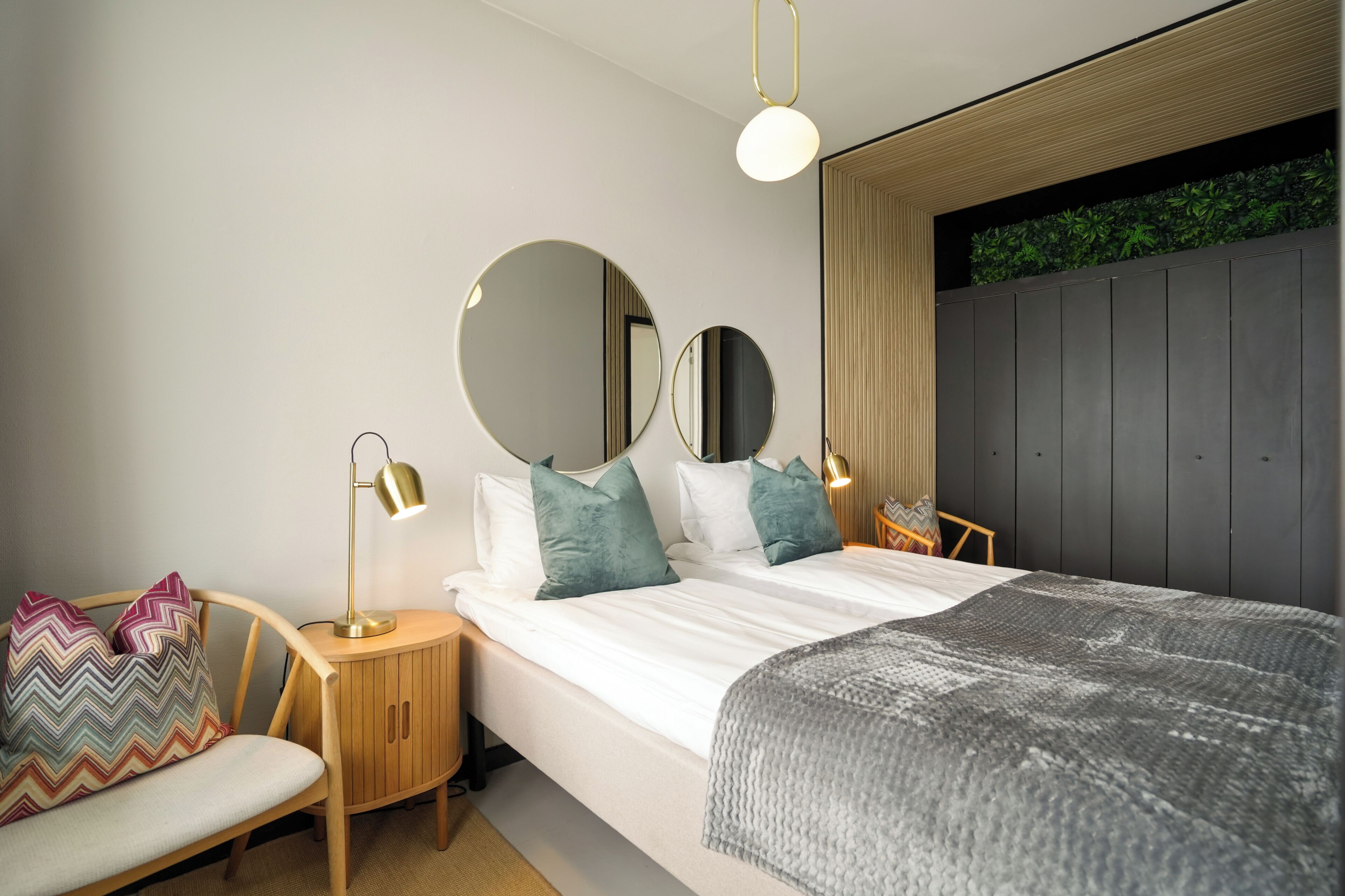 Quarto moderno com cama, duas mesas de cabeceira, uma cadeira, espelho redondo e planta suspensa na parede.