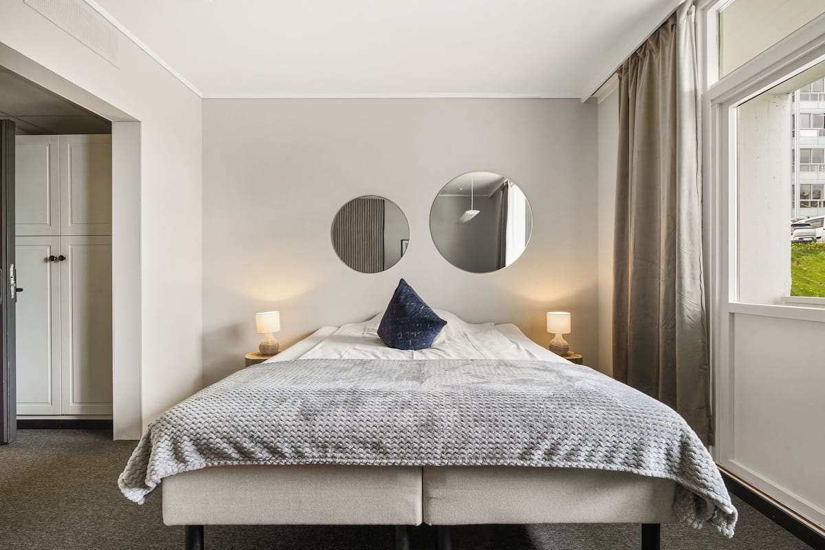 A bedroom with a bed, two bedside lamps, a window with curtains, and a door to another room.