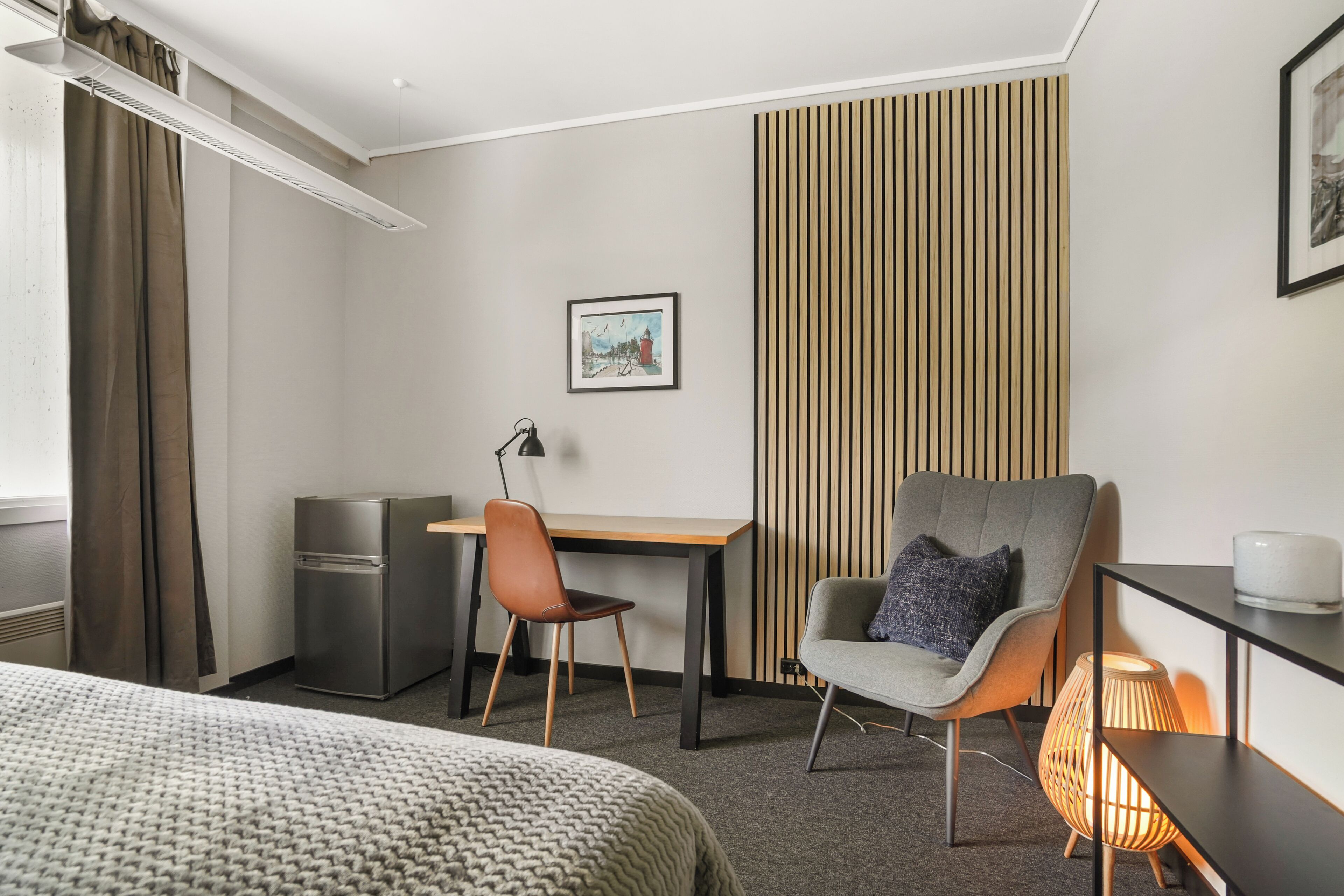 A hotel room with a bed, a desk, a chair, a small refrigerator, a lamp, a picture on the wall, and a bedside table with a lamp.