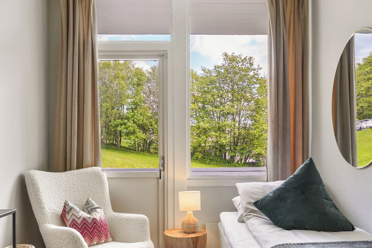 A hotel room with a bed, a chair, a small table with a lamp, and a view of a green landscape through a window.