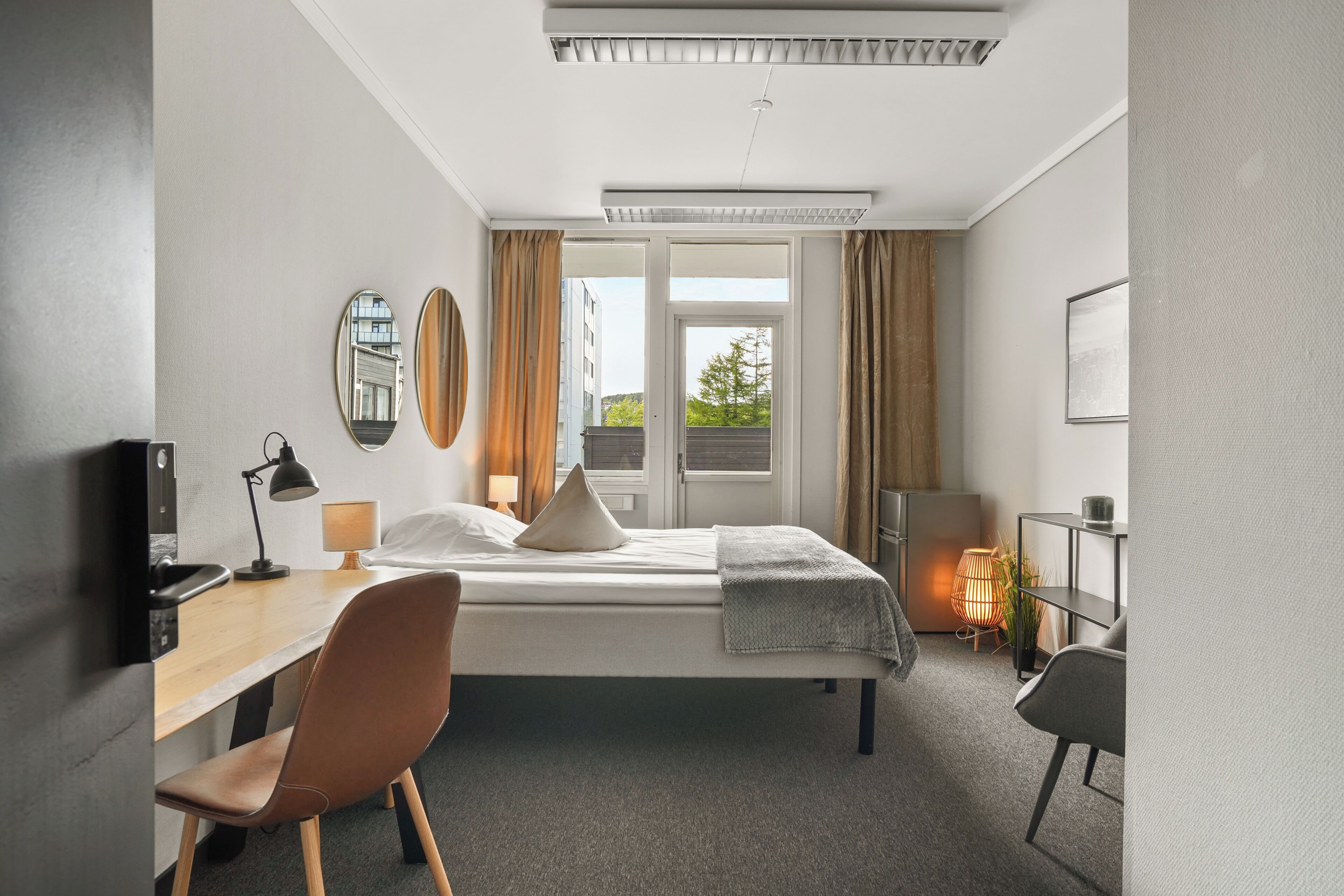 Une chambre d’hôtel moderne équipée d’un lit, d’un bureau, d’une chaise et d’une grande fenêtre avec des rideaux.