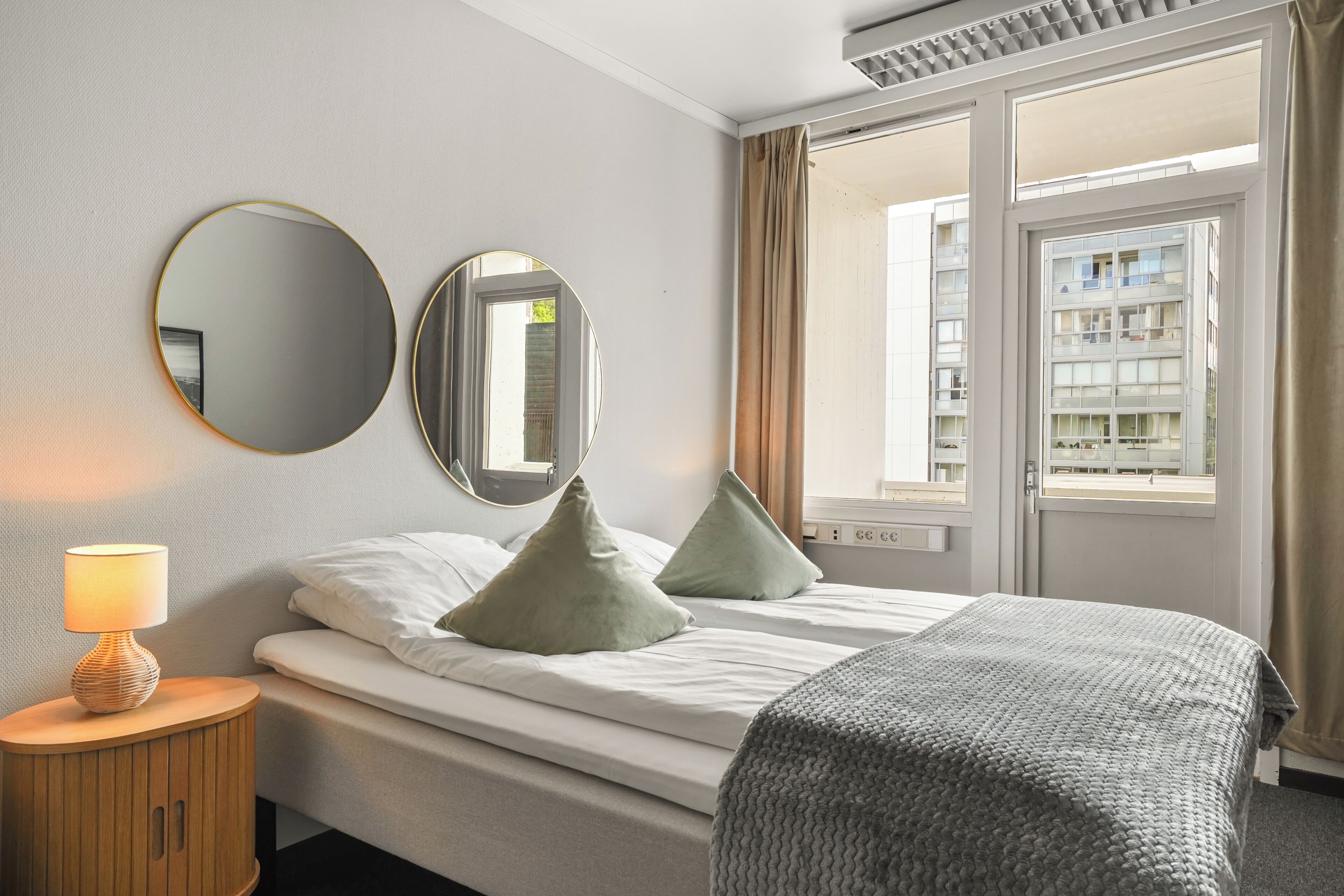 Habitación de hotel con cama, dos espejos a los lados, lámpara y ventana con cortinas.