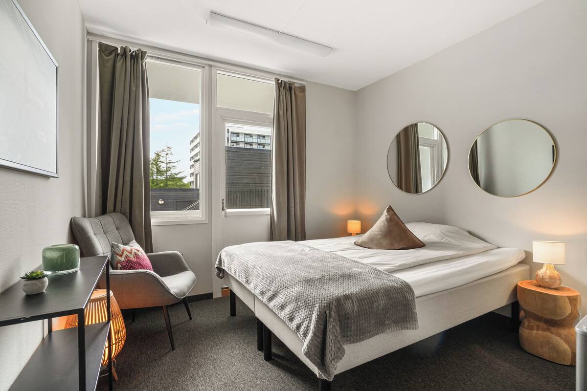 A hotel room with a bed, a chair, a desk, a lamp, and two mirrors.