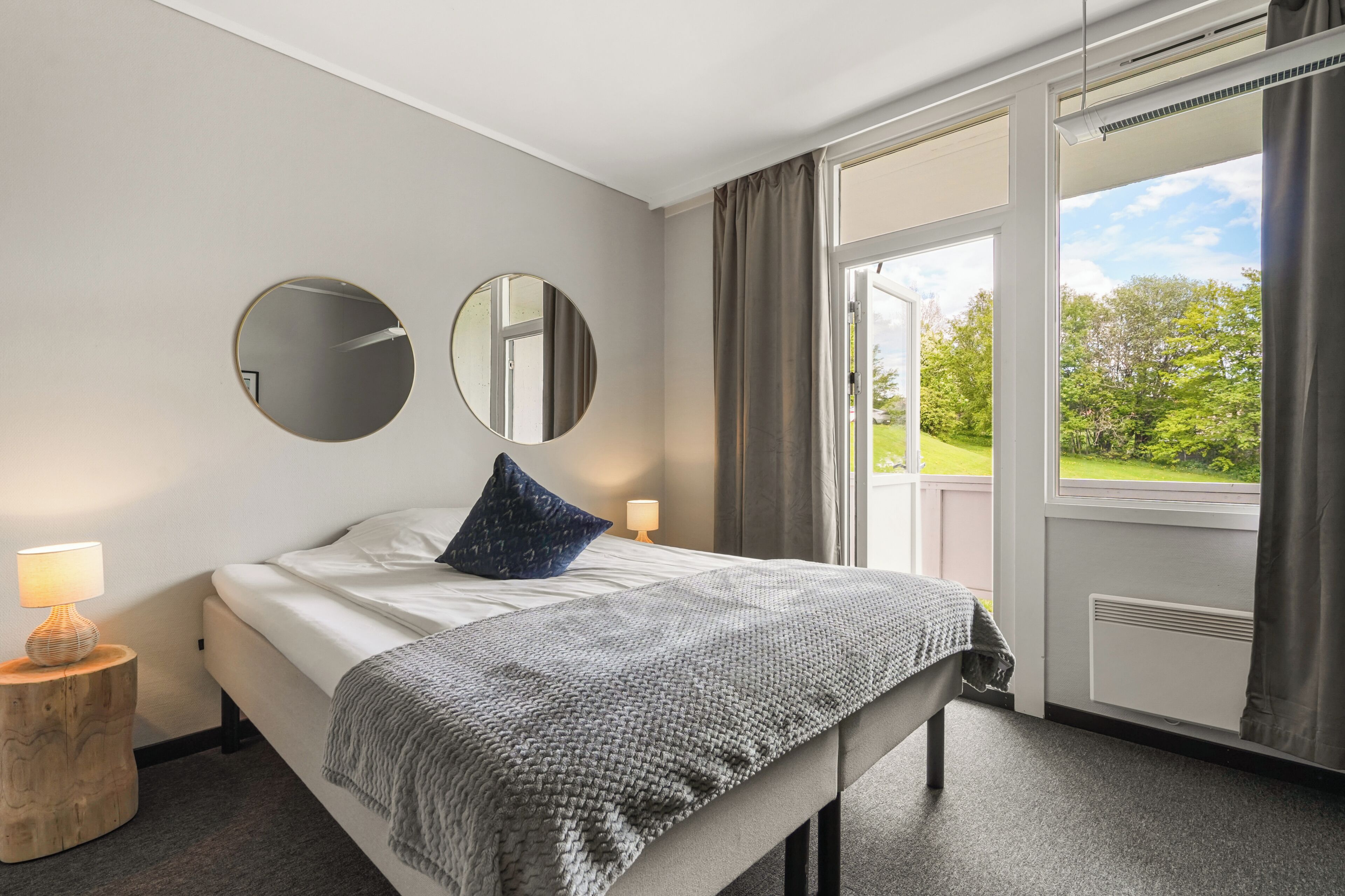 A hotel room with a bed, two bedside lamps, a round mirror, a window with curtains, and a view of greenery outside.