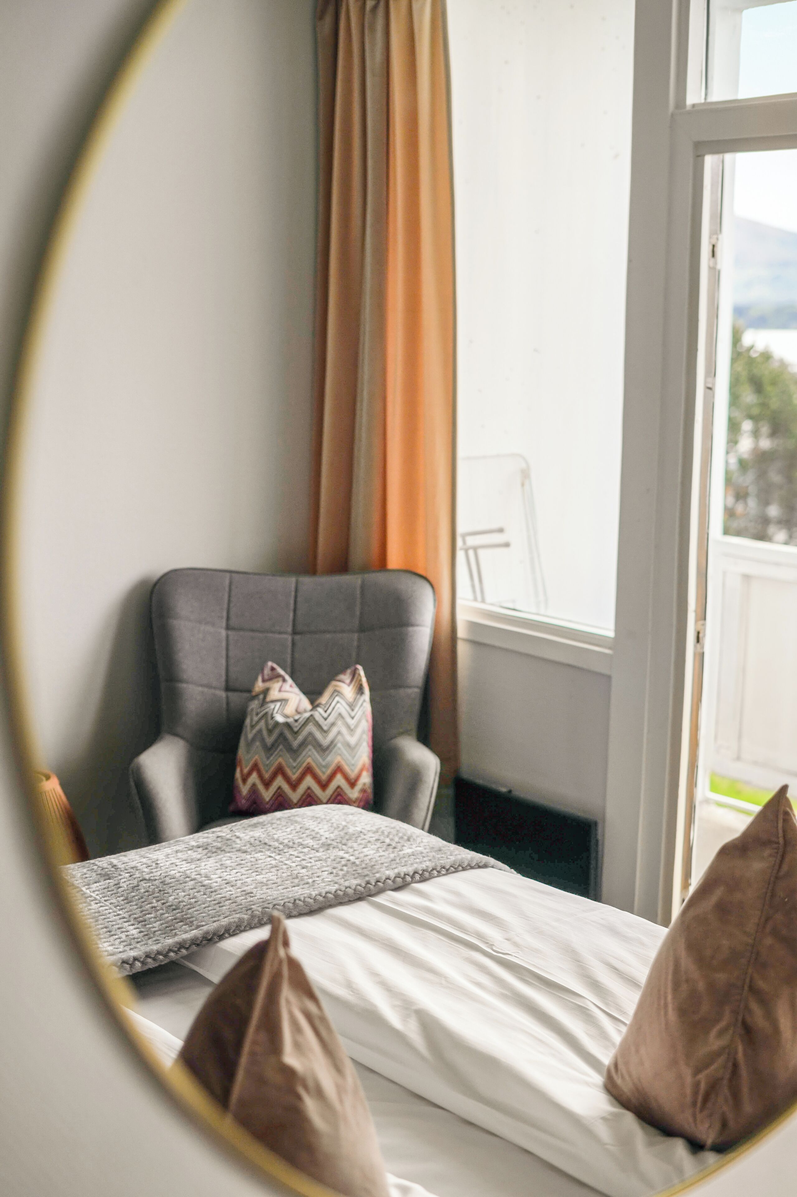 Een kamer met een bed, een grijze fauteuil, een spiegel en een raam met gordijnen.