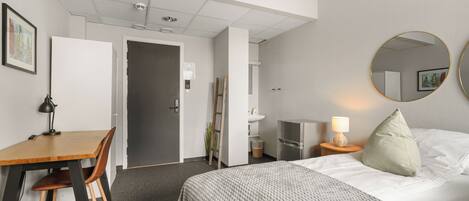 A hotel room with a bed, a desk, a chair, a TV, a lamp, a mirror, and a door leading to another room.