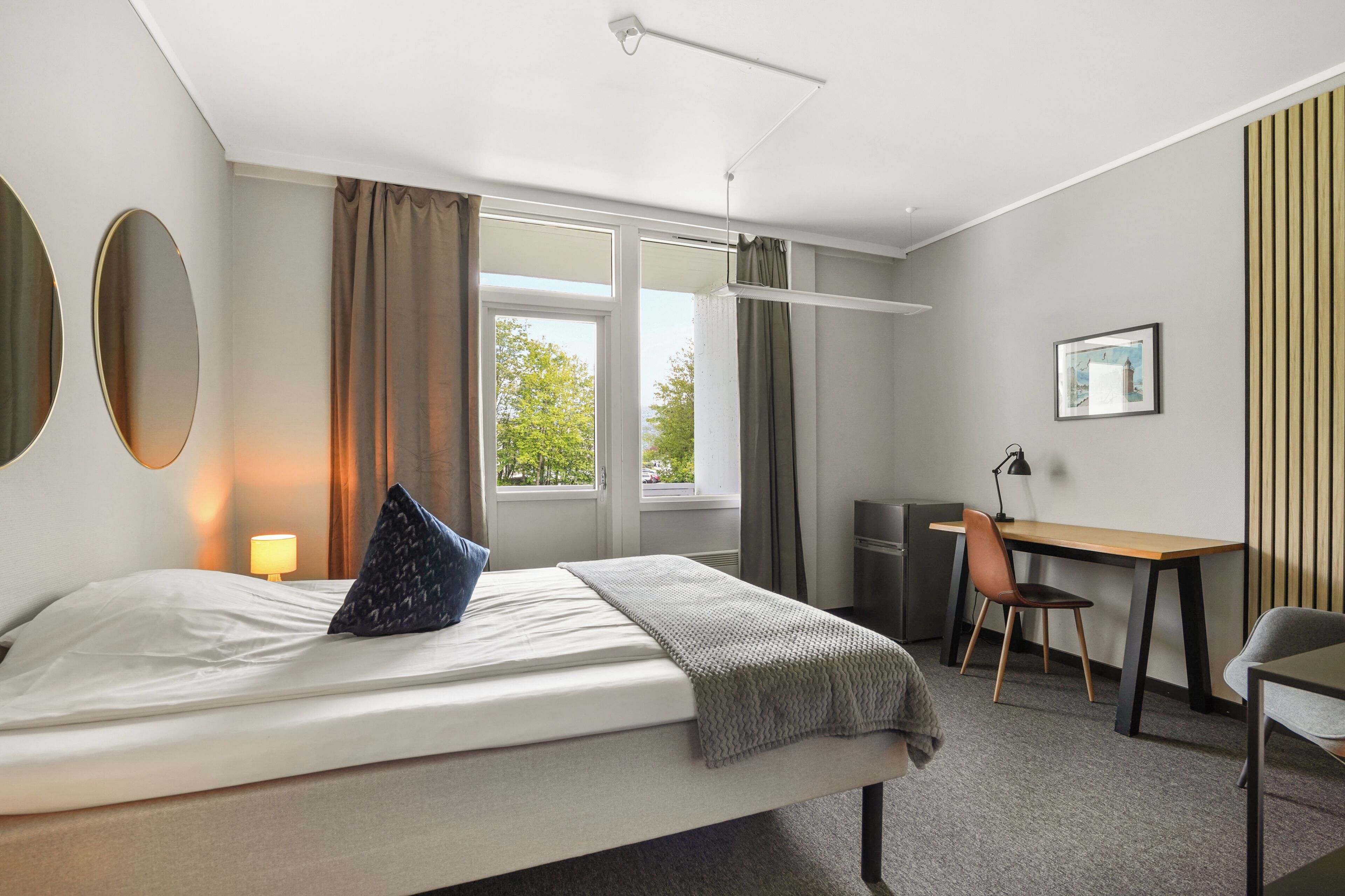 A hotel room with a bed, desk, chair, and a window with curtains.