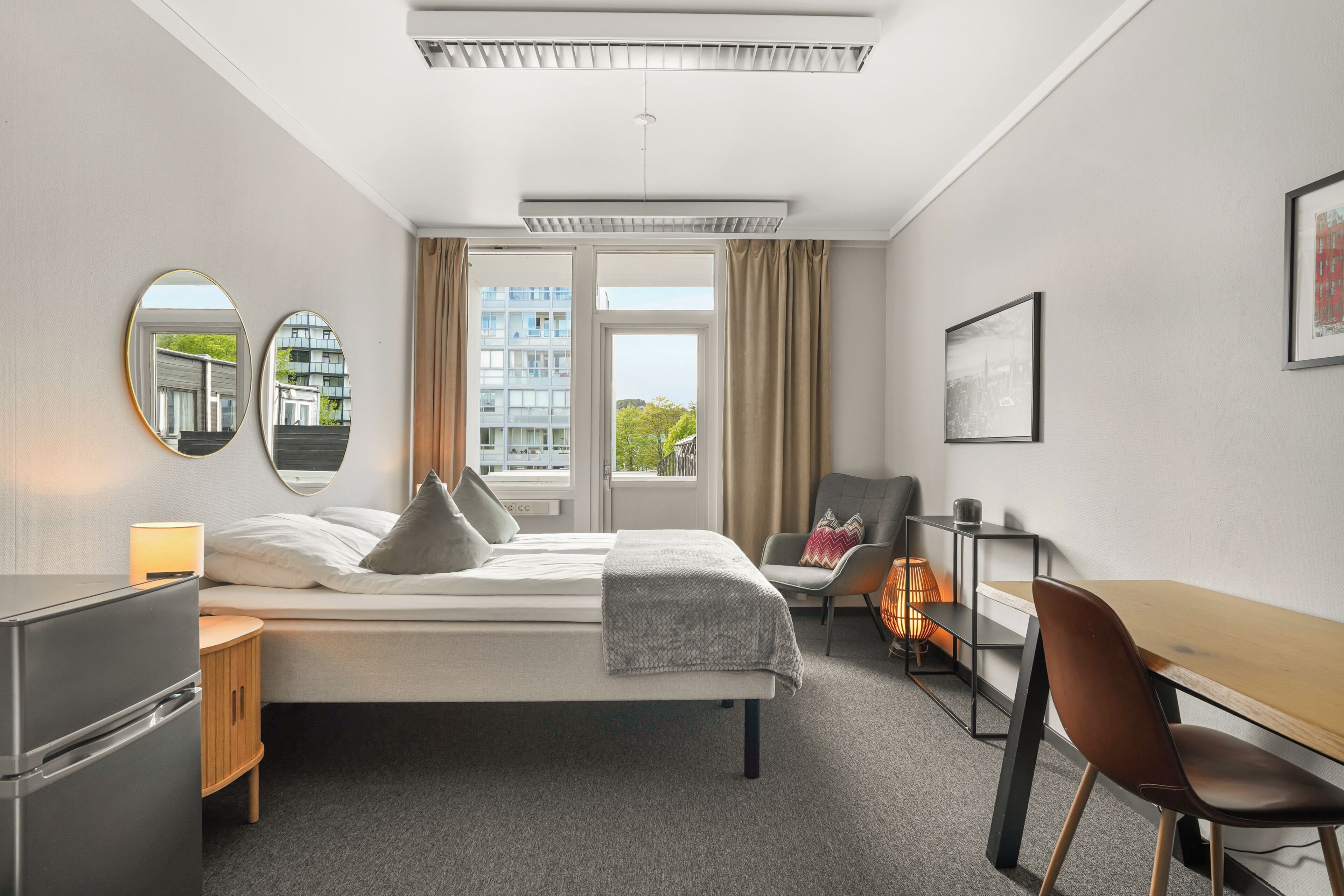 A hotel room with a bed, a desk, a chair, a lamp, a mirror, and a window with curtains.