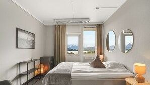 A hotel room with a bed, a desk, a chair, a lamp, a shelf, and a window with a view of mountains. - A-11 (Ålesund)