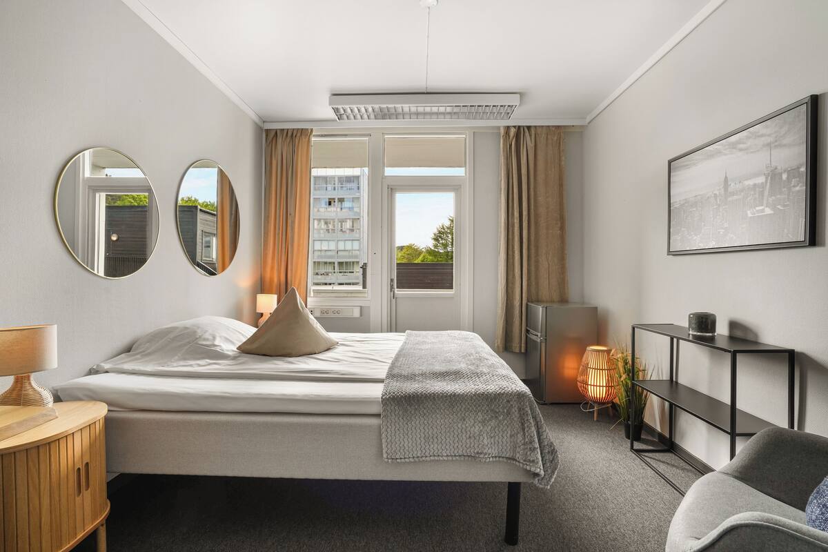 A modern hotel room with a bed, bedside tables, a desk, a chair, a TV, a bookshelf, and a window with curtains.