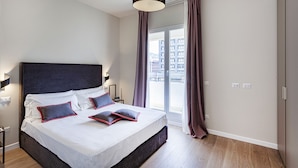 Quarto com uma cama grande, janela com cortinas, luminária pendente e vista para edifícios.