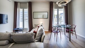 Uma sala de estar moderna com sofá, mesa de jantar, cadeiras, televisão de tela plana e uma grande janela com cortinas.