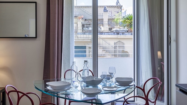 Uma mesa de jantar com tampo de vidro e cadeiras de metal, uma janela com cortinas, e vista para edifícios e uma estátua lá fora.