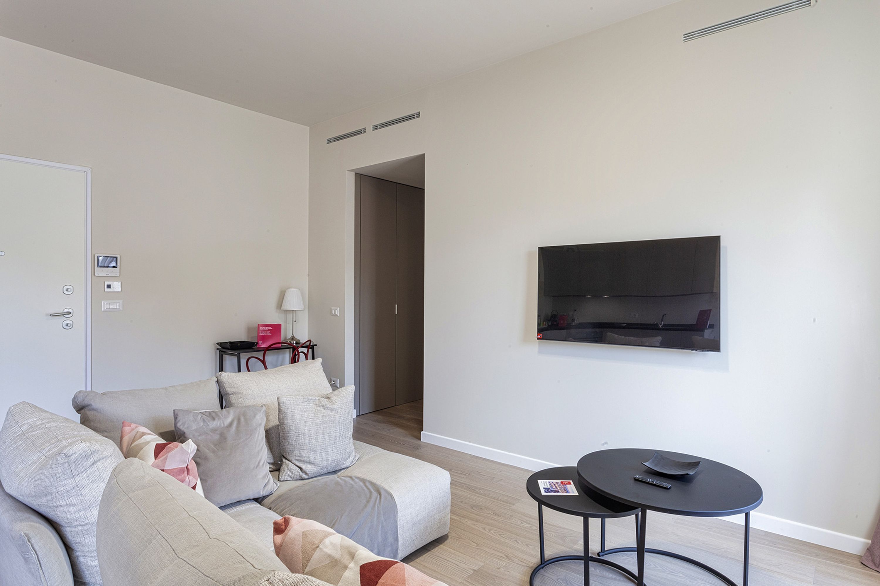 A modern living room with a sofa, a small round table, a TV mounted on the wall, and a door with a lock.