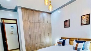 A bedroom with a large bed, wooden wardrobe, and a ceiling light fixture. - Eden Homes (Kampala)