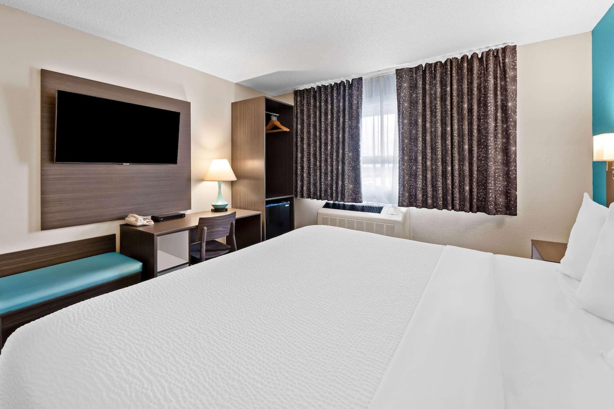 Habitación de hotel con una cama grande, un escritorio con lámpara, un televisor en la pared y una ventana con cortinas.