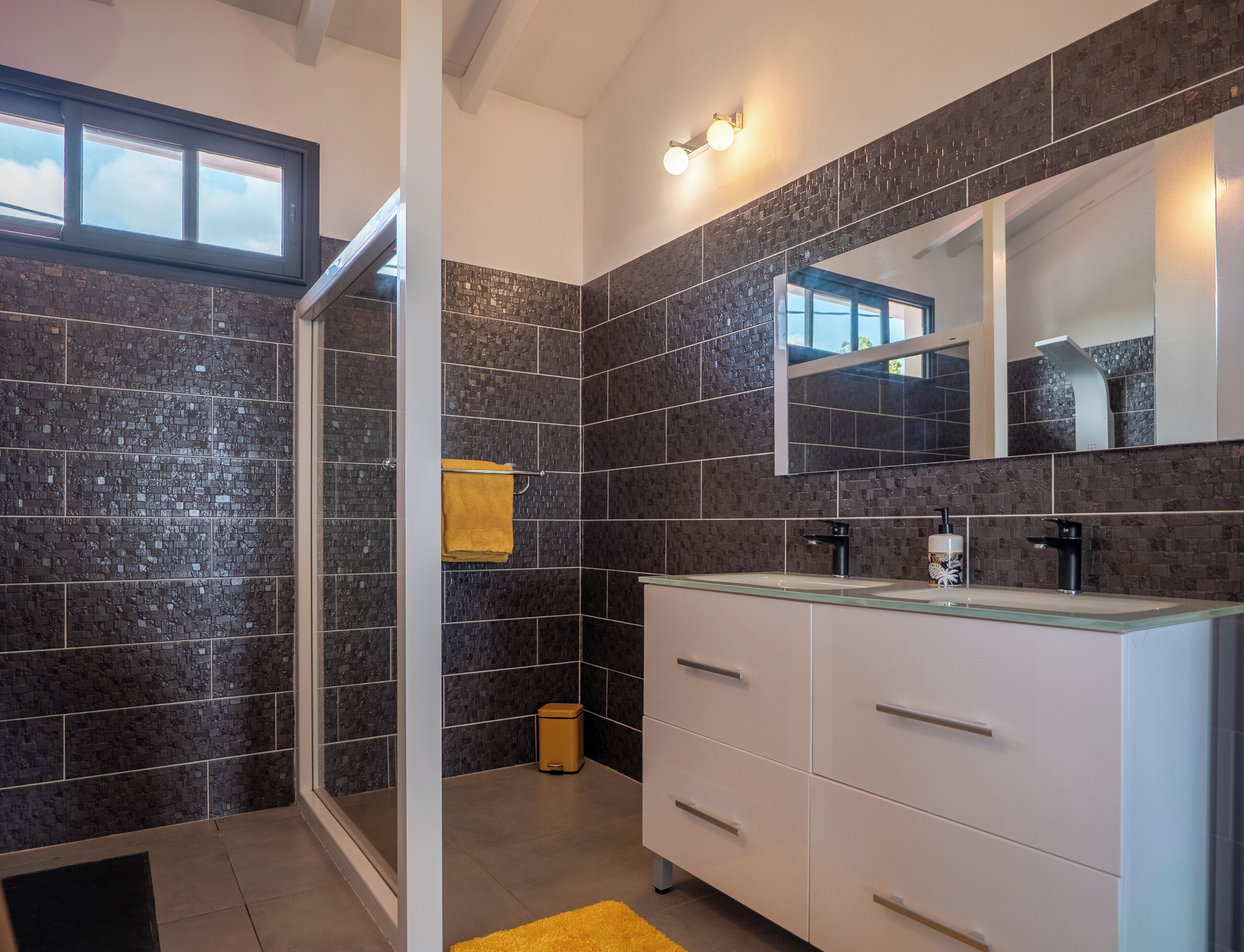 A modern bathroom with dark tiled walls, a glass shower enclosure, white cabinetry, and a yellow rug.