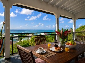 A wooden table with breakfast items, a view of the ocean, and a balcony with a railing. - Penthouse Luxe • Vue Mer Panoramique 180° • Terrasse • Plage à pied (Sainte-Anne)
