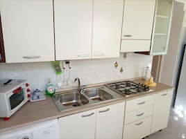 Una cocina con armarios blancos, un lavamanos de acero inoxidable, estufa a gas, microondas y escurreplatos.