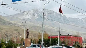 A street scene with a statue, a red building, and cars parked on the side of the road. - DADOW Apartments (Naryn)