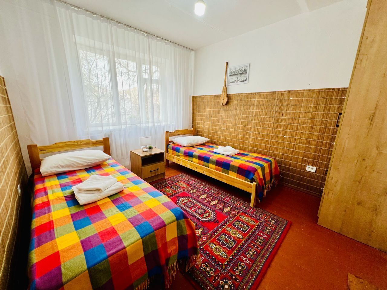 Habitación con dos camas, cabecera de madera, mesita de noche pequeña, estante en la pared y alfombra estampada.