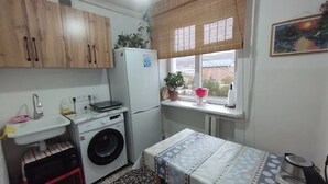 A compact kitchen with wooden cabinets, a washing machine, a sink, a window with a view, and a table with a patterned tablecloth. - DADOW Apartments (Naryn)