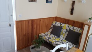A corner room with a wooden panel wall, a patterned sofa, a small table with a plant, and a chair. - Villa Géraniums Apt n°4 (Mont-Dore)