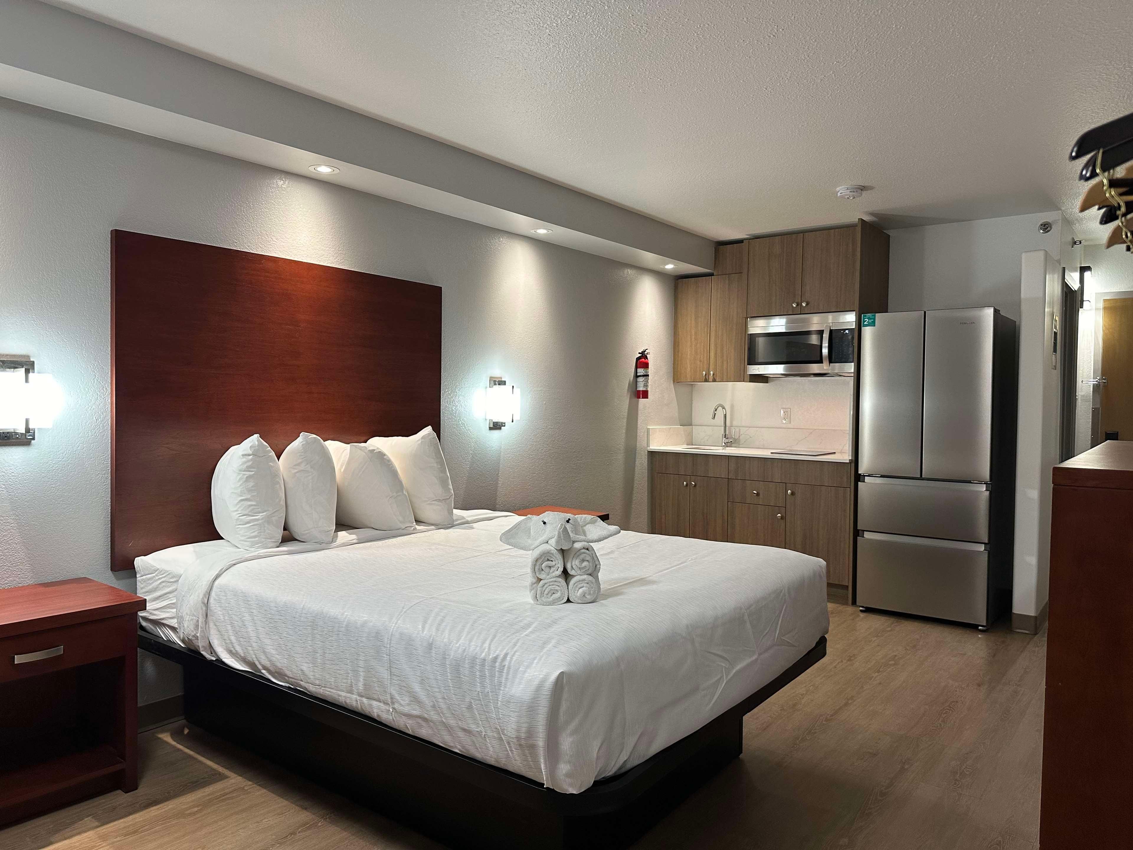 Une chambre d’hôtel comprenant un lit, une table de chevet, une lampe fixée au mur, une tête de lit en bois, une cuisine intégrée avec micro-ondes et réfrigérateur, et un extincteur.