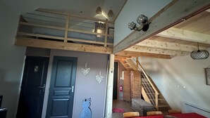 A loft-style bedroom with a high ceiling, exposed wooden beams, a red bed, and a staircase leading to an upper level.