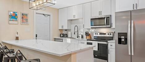 A modern kitchen with white cabinets, a large island with bar stools, a stainless steel refrigerator, and a built-in microwave.
