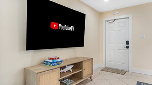 Una habitación con un televisor de pantalla plana montado en la pared que muestra el logotipo de YouTubeTV, una mesita de madera con una figura de tiburón y una pila de cajas, y una puerta blanca con felpudo.