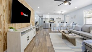 A modern living room with a flat-screen TV, a ceiling fan, and a kitchen area with white cabinets and a dining set. - The Shark Tank: Coastal Modern, Walk to Beach, Shops & Downtown Cocoa Beach (Cocoa Beach)