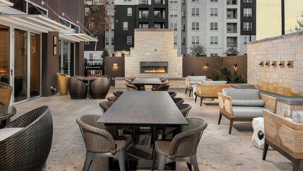 Une terrasse extérieure dotée d’une cheminée en pierre, de meubles en osier et offrant une vue sur des immeubles d’habitation.