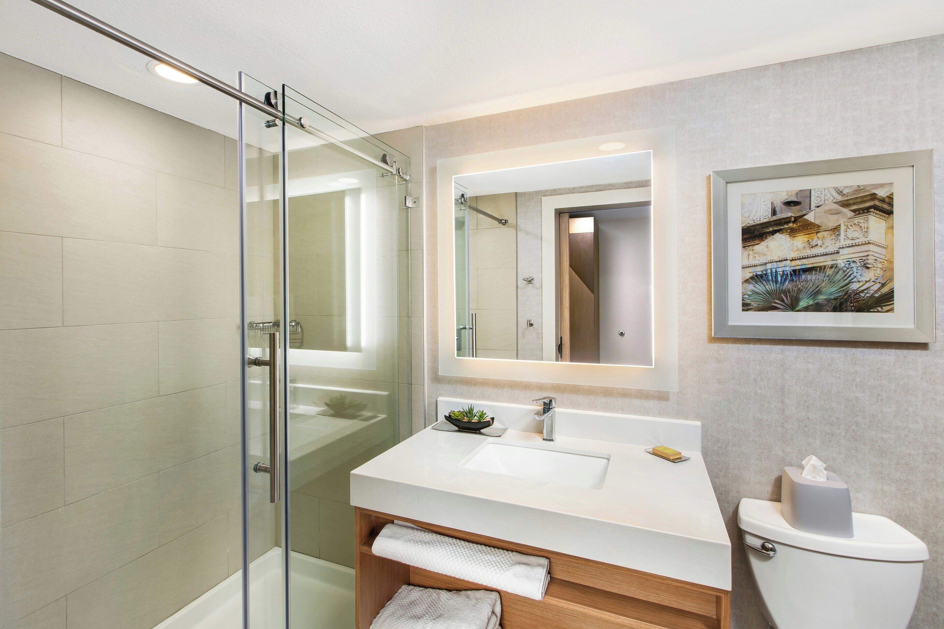 A modern bathroom with a glass shower enclosure, a white sink with a mirror, and a framed artwork on the wall.