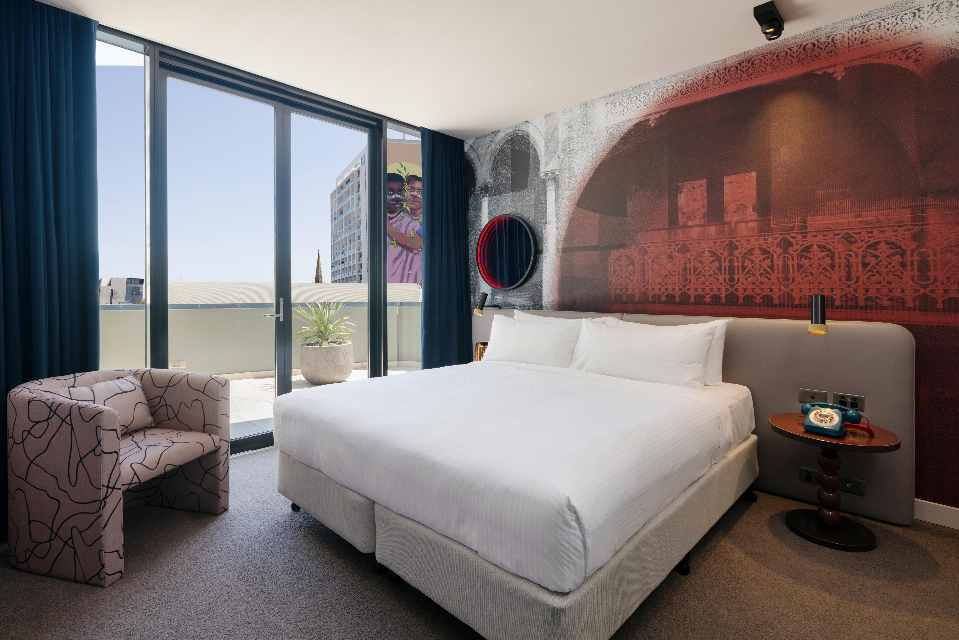 A hotel room with a large bed, a chair, a small table with a telephone, and a view of the city through a large window.