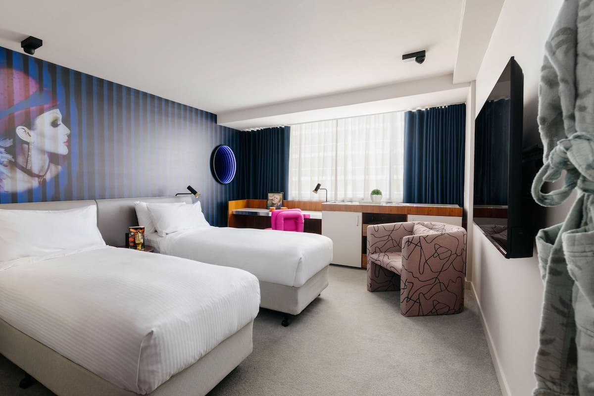 A hotel room with two beds, a desk, a chair, a wall-mounted lamp, a small table with a plant, and a large mirror.