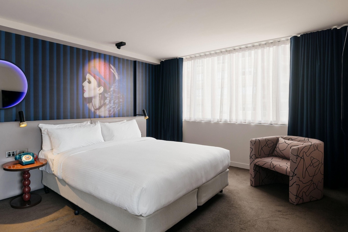 Una habitación de hotel con cama, un espejo redondo, una mesita con teléfono, una silla y un amplio ventanal con cortinas.