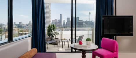 Ein modernes Hotelzimmer mit großem Balkon, einer Couch, einem rosa Sessel, einem kleinen Tisch mit einem Buch und einem Fernseher.