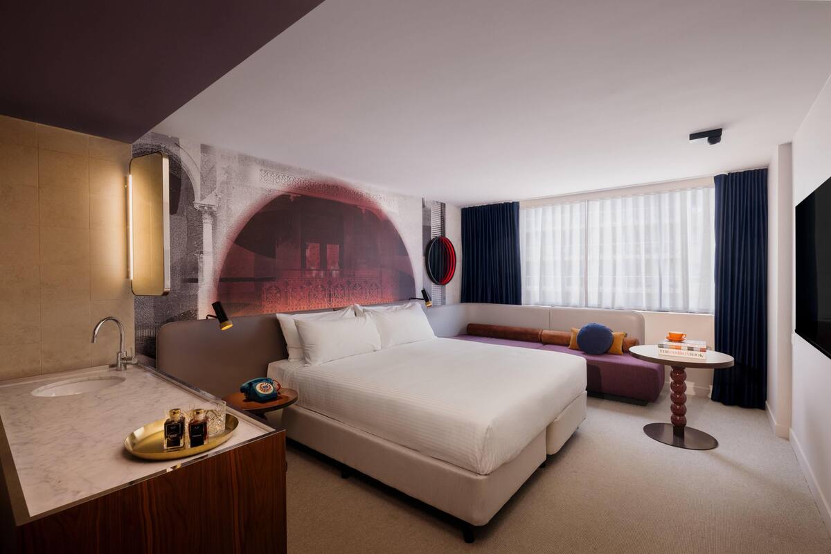 A hotel room with a bed, a sofa, a small table, a sink, and a mirror.