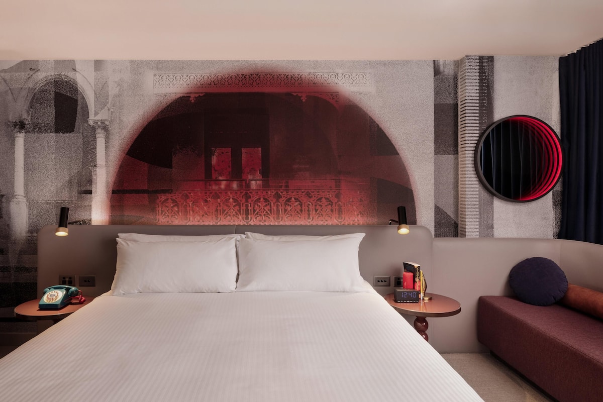 A hotel room with a bed, a sofa, a round mirror, a telephone, and a small table with a clock.
