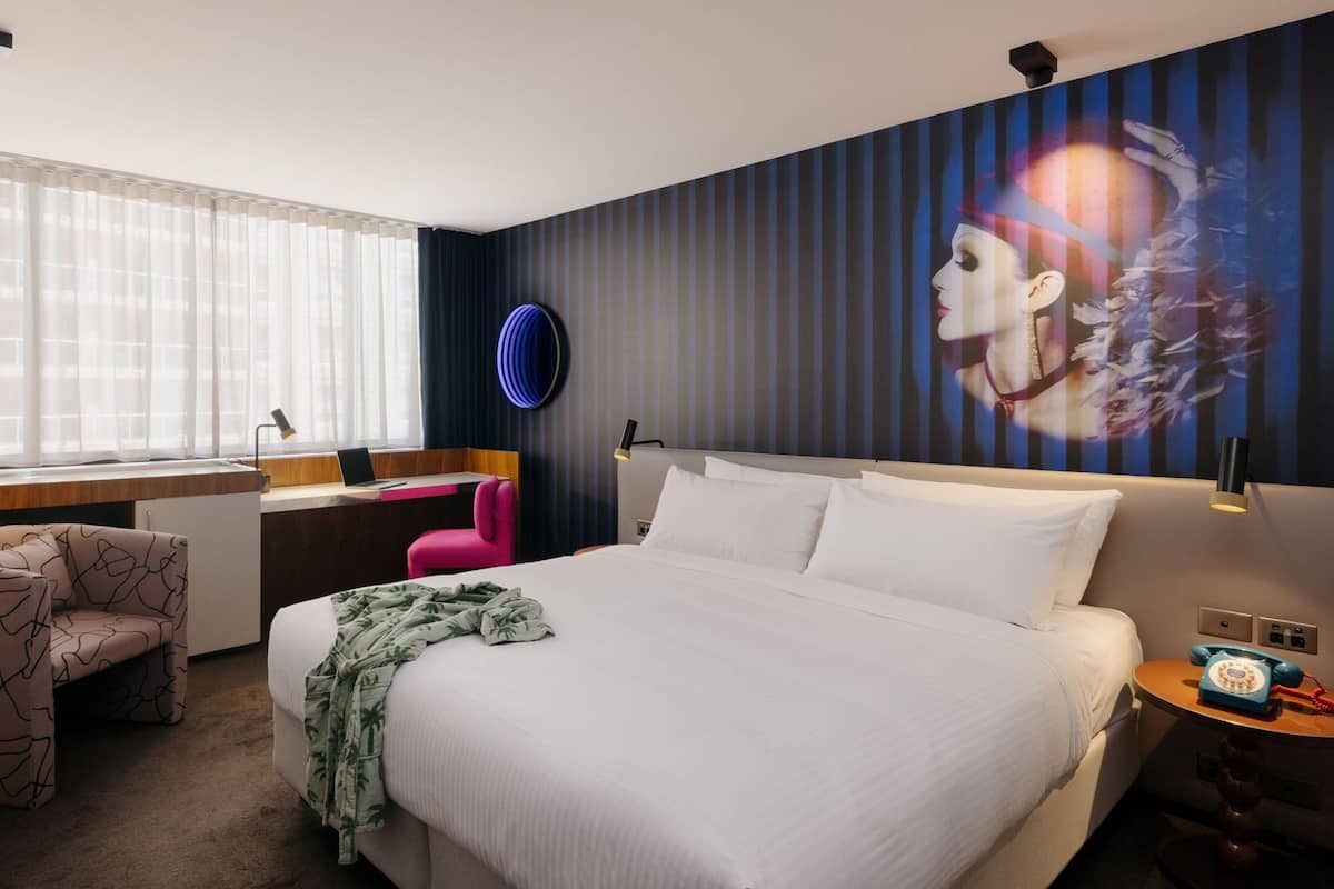 A hotel room with a bed, a desk, a chair, a phone, and a wall with a mural.